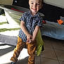 child, boy, smile, indoor, sunlight, shadow, checkered_shirt, mustard_pants, black_shoes, tile_floor, bed, blanket, laundry_basket, hair_spiky, happy, casual_clothing, home, person, floor, playful