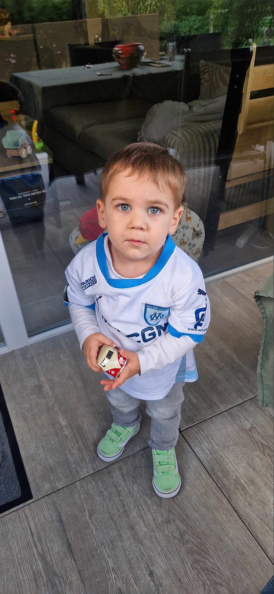 Nolann participe au concours pour gagner de l'argent avec cette photo : child, toddler, indoor, toy, holding, blue_eyes, sneakers, shirt, floor, reflection, glass, sofa, chair, curious, standing, casual_clothing, young, person, looking_up, home