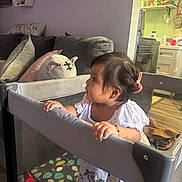 Nazneen is registered to the contest to win money with this photo: toddler, child, playpen, white_dress, bracelet, living_room, couch, pillows, toy, mesh, curly_hair, earrings, home_interior, kitchen, cabinet, floor, furniture, person, indoor, standing