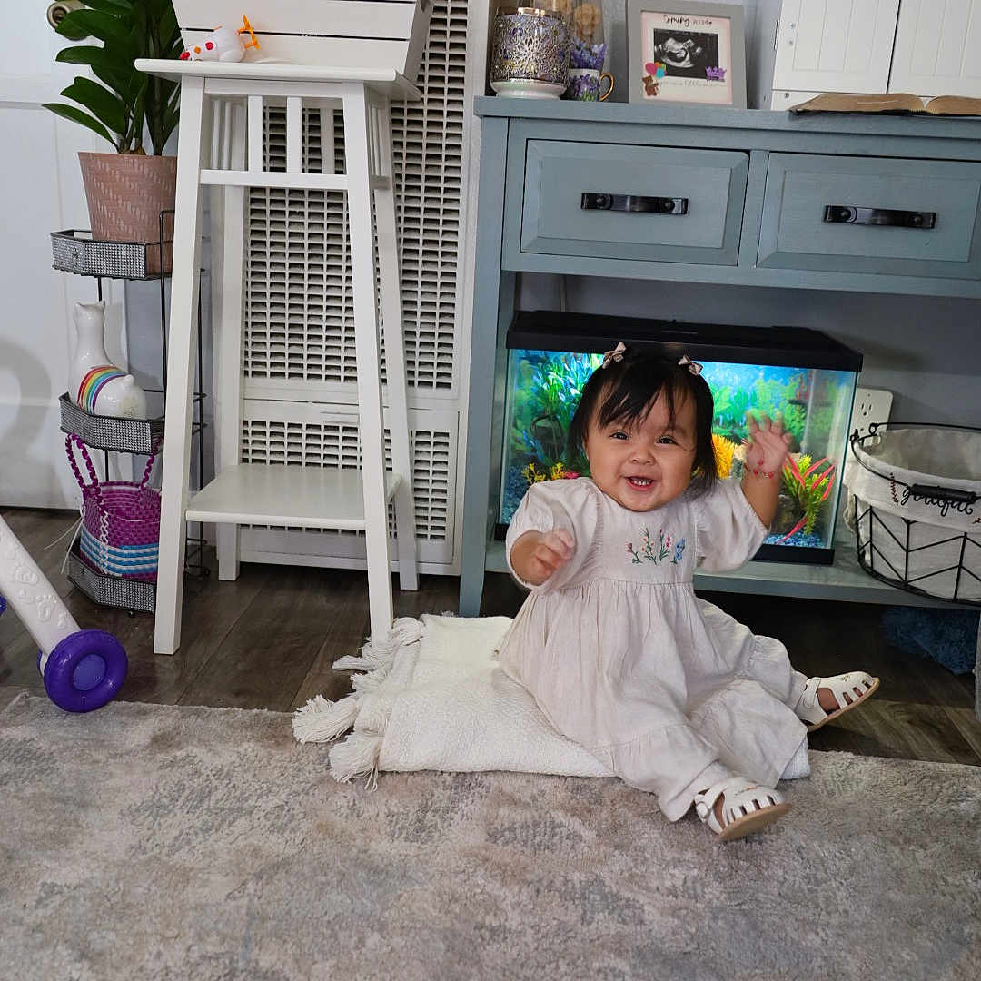 Nazneen joined the competition — help win amazing prizes! aquarium, baby, basket, blanket, cabinet, carpet, child, decor, dress, flower_pot, furniture, happy, indoor, living_room, person, plant, smiling, teddy_bear, toy, wooden_floor