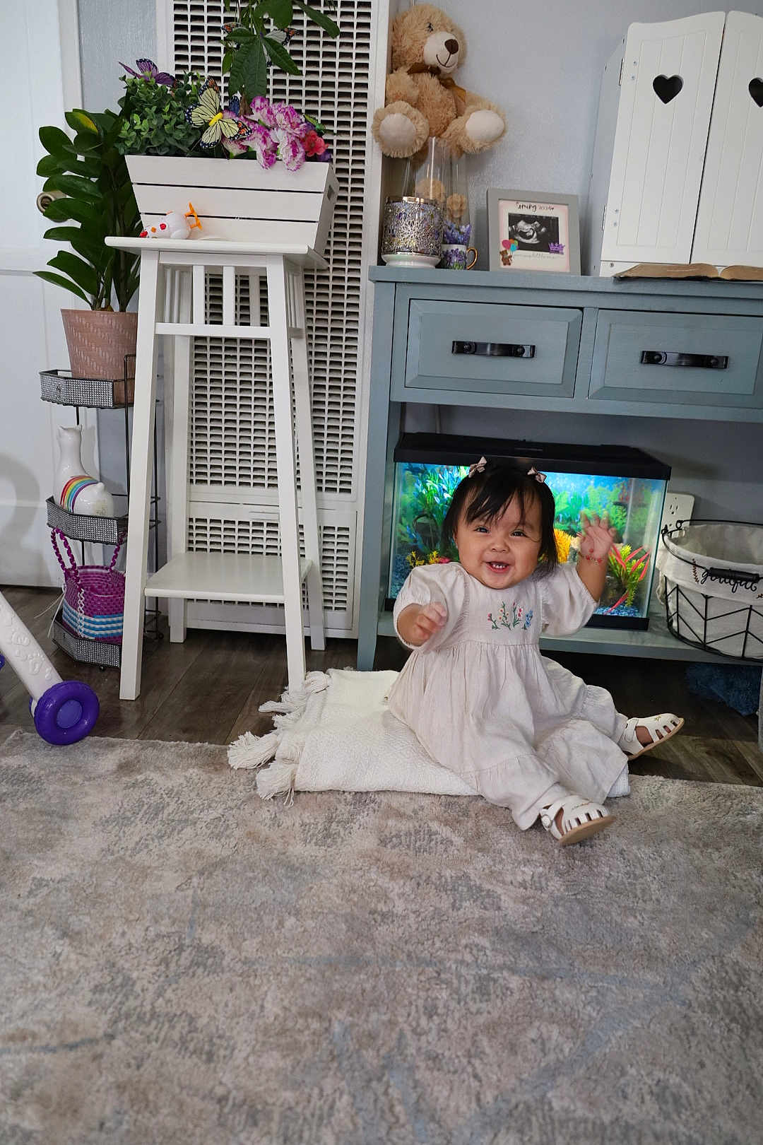 Nazneen joined the competition — help win amazing prizes! baby, child, smiling, indoor, dress, blanket, carpet, furniture, cabinet, aquarium, teddy_bear, plant, flower_pot, toy, wooden_floor, decor, basket, happy, person, living_room