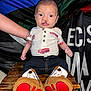 Daxen is registered to the contest to win money with this photo: baby, child, infant, cleft_lip, cowboy_boots, shirt, wooden_stool, arm, flag, text, wide_eyes, denim_pants, indoor, person, footwear, seated, background, casual_clothing, close_up, portrait