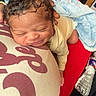 newborn, baby, pillow, blanket, infant, sleeping, resting, hand, red_clothing, water_bottle, curly_hair, indoors, soft_texture, peaceful, closeup, person, candid, comfort, head, skin