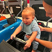Auziel is registered to the contest to win money with this photo: adult, baby, background, bib, black_car, blue_car, child, floor, foot, happy, holding, indoor, person, playful, seat, smiling, steering_wheel, striped_clothing, supporting_hand, toy_car