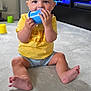 baby, child, sitting, carpet, indoor, toy, plastic_cup, yellow_shirt, blue_shorts, barefoot, curious, cute, living_room, furniture, candle, television, window, curtain, floor, home