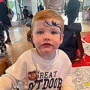 Bertie is registered to the contest to win money with this photo: toddler, child, face_paint, bat, indoor, table, coloring_sheets, marker, white_sweatshirt, people, event, backpack, chair, window, curtains, wooden_floor, casual_clothing, portrait, expression, holiday