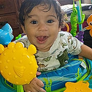 Kai is registered to the contest to win money with this photo: toddler, child, smiling, happy, toy, indoor, playtime, curly_hair, face, eyes, clothing, baby_seat, yellow_toy, blue_toy, orange_toy, window, wooden_cabinet, person, expression, cute