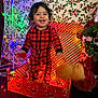 background, celebration, child, christmas, christmas_lights, curly_hair, decorations, face, festive, gift_box, happy, holiday_decor, indoor, joyful, necklace, plaid, red_pajamas, smiling, toddler, toy