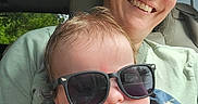 Ronin is registered to the contest to win money with this photo: baby, child, mother, adult, sunglasses, smiling, pouting, portrait, selfie, car, vehicle, seated, family, parent, infant, blue_shirt, green_shirt, indoors, daylight, photography