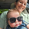baby, child, mother, adult, sunglasses, smiling, pouting, portrait, selfie, car, vehicle, seated, family, parent, infant, blue_shirt, green_shirt, indoors, daylight, photography