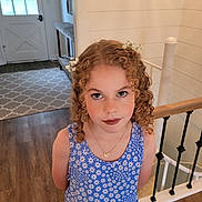 Scarlett is registered to the contest to win money with this photo: child, girl, portrait, curly_hair, flower_crown, blue_dress, floral_pattern, smiling, indoors, home, staircase, wooden_floor, railing, entryway, door, window, necklace, standing, face, photography
