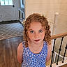 child, girl, portrait, curly_hair, flower_crown, blue_dress, floral_pattern, smiling, indoors, home, staircase, wooden_floor, railing, entryway, door, window, necklace, standing, face, photography