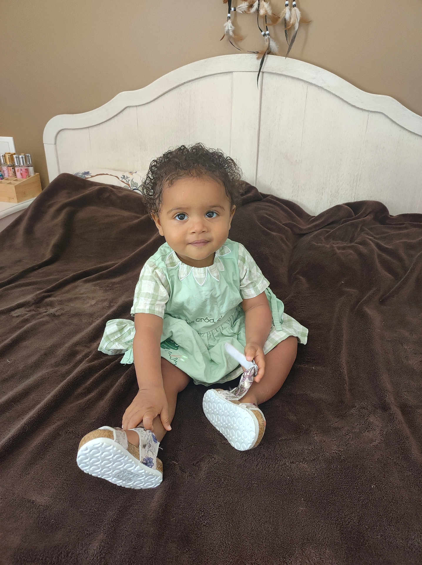 Malika participe au concours pour gagner de l'argent avec cette photo : baby, child, toddler, curly_hair, green_dress, shoes, bed, blanket, brown_blanket, headboard, indoor, portrait, smiling, sitting, furniture, person, cute, soft_lighting, looking_at_camera, home