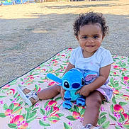 Malika a rejoint le concours — aidez-le/la à gagner de superbes lots ! child, toddler, curly_hair, plush_toy, blue_toy, blanket, floral_pattern, outdoor, park, grass, summer, shoes, cute, smiling, sitting, playtime, nature, sunlight, daytime, happy
