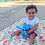 child, toddler, curly_hair, plush_toy, blue_toy, blanket, floral_pattern, outdoor, park, grass, summer, shoes, cute, smiling, sitting, playtime, nature, sunlight, daytime, happy
