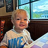 toddler, child, smiling, portrait, indoor, restaurant, table, checkered_tablecloth, window, mountains, scenic_view, blond_hair, blue_eyes, nike_shirt, wood_paneling, chair, framed_pictures, daylight, happy, candid