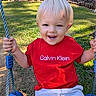 toddler, child, smiling, swing, playground, outdoor, backyard, grass, red_shirt, white_pants, blond_hair, portrait, happy, sitting, ropes, sunlight, garden, play, face, clothing