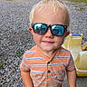 child, toddler, sunglasses, portrait, outdoor, smiling, blond_hair, striped_shirt, orange_shirt, face, person, gravel, backyard, playground, chair, pool, trailer, trees, reflection, photography