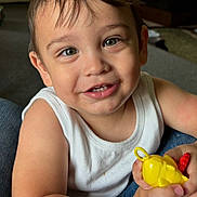 Ronnie joined the competition — help win amazing prizes! toddler, child, smiling, indoor, toy, yellow_toy, white_tank_top, happy, portrait, cute, close_up, person, skin, hair, eyes, furniture, carpet, home, casual, sitting