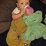 Cyrus is registered to the contest to win money with this photo: baby, child, bottle, overalls, plush_toy, frog, toy, couch, indoor, person, cute, smiling, hand, foot, clothing, fabric, seat, comfort, infant, drink