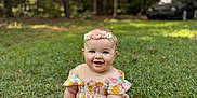 Aria is registered to the contest to win money with this photo: baby, child, grass, outdoor, smiling, flower_headband, dress, greenery, nature, sunlight, trees, parked_car, cute, happy, sitting, portrait, infant, summer, playful, young