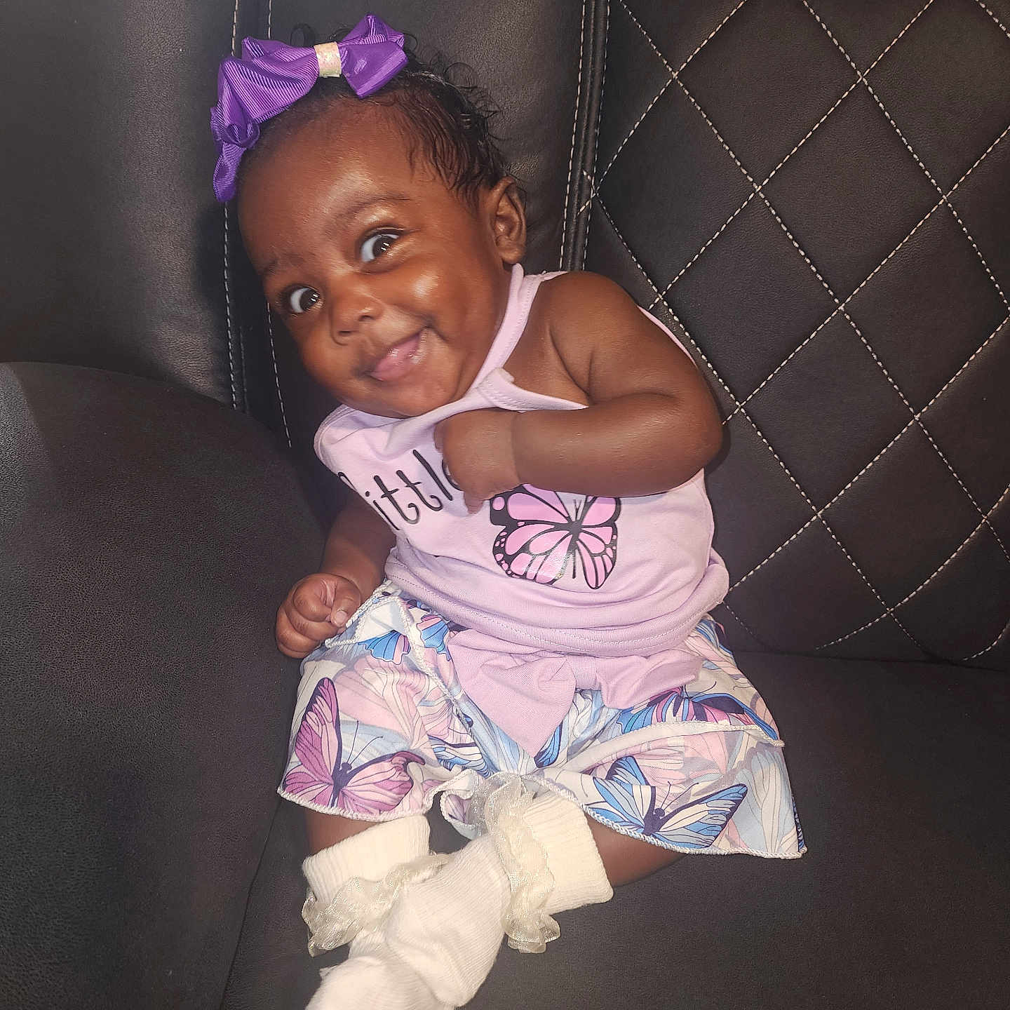Jakerya is registered to the contest to win money with this photo: baby, child, smiling, purple_bow, butterfly, dress, socks, couch, leather, quilted, happy, cute, infant, portrait, indoor, person, seated, clothing, face, skin