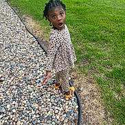 Baila joined the competition — help win amazing prizes! child, girl, braided_hair, leopard_print, outdoor, grass, stones, rocks, sidewalk, shoes, surprised_expression, standing, nature, casual_clothing, young_child, curious, daylight, playful, person, greenery