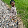 child, girl, braided_hair, leopard_print, outdoor, grass, stones, rocks, sidewalk, shoes, surprised_expression, standing, nature, casual_clothing, young_child, curious, daylight, playful, person, greenery