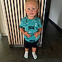 blonde_hair, blue_eyes, casual_clothing, child, curious, cute, floor, home, indoor, modern_interior, person, portrait, shorts, socks, standing, toddler, tshirt, wall, wooden_panel, young_child