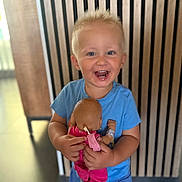Ezio a rejoint le concours — aidez-le/la à gagner de superbes lots ! blonde_hair, blue_shirt, casual_clothing, child, cute, doll, face, happy, holding, indoor, joyful, person, pink_clothing, playful, short_hair, small_child, smiling, toddler, toy, wooden_slats