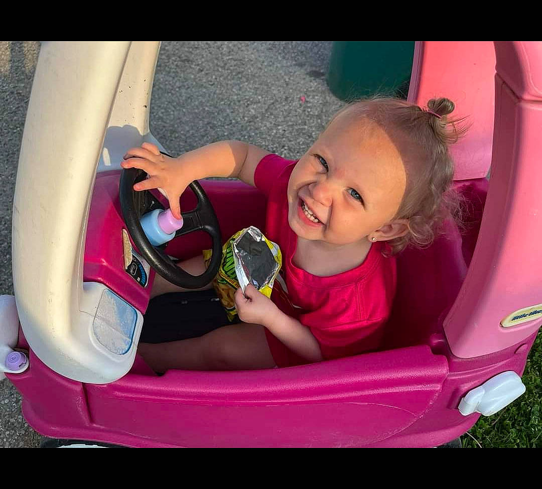 Brylie is registered to the contest to win money with this photo: arm, automotive_design, automotive_exterior, automotive_tire, baby_carriage, bumper, car, fender, hand, happy, joy, motor_vehicle, person, pink, plant, product, smile, tire, toddler, vehicle