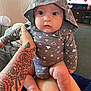 baby, carpet, casual, comfort, cute, foot, furniture, hand, hoodie, indoor, infant, legs, living_room, patterned_clothing, person, sitting, skin, tattoo, television, wide_eyes