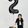toddler, child, balloon, number_two, gift_box, black_clothing, white_shoes, studio, portrait, celebration, present, ribbon, standing, casual_wear, expressionless, indoor, minimalist, curly_hair, fashion, isolated