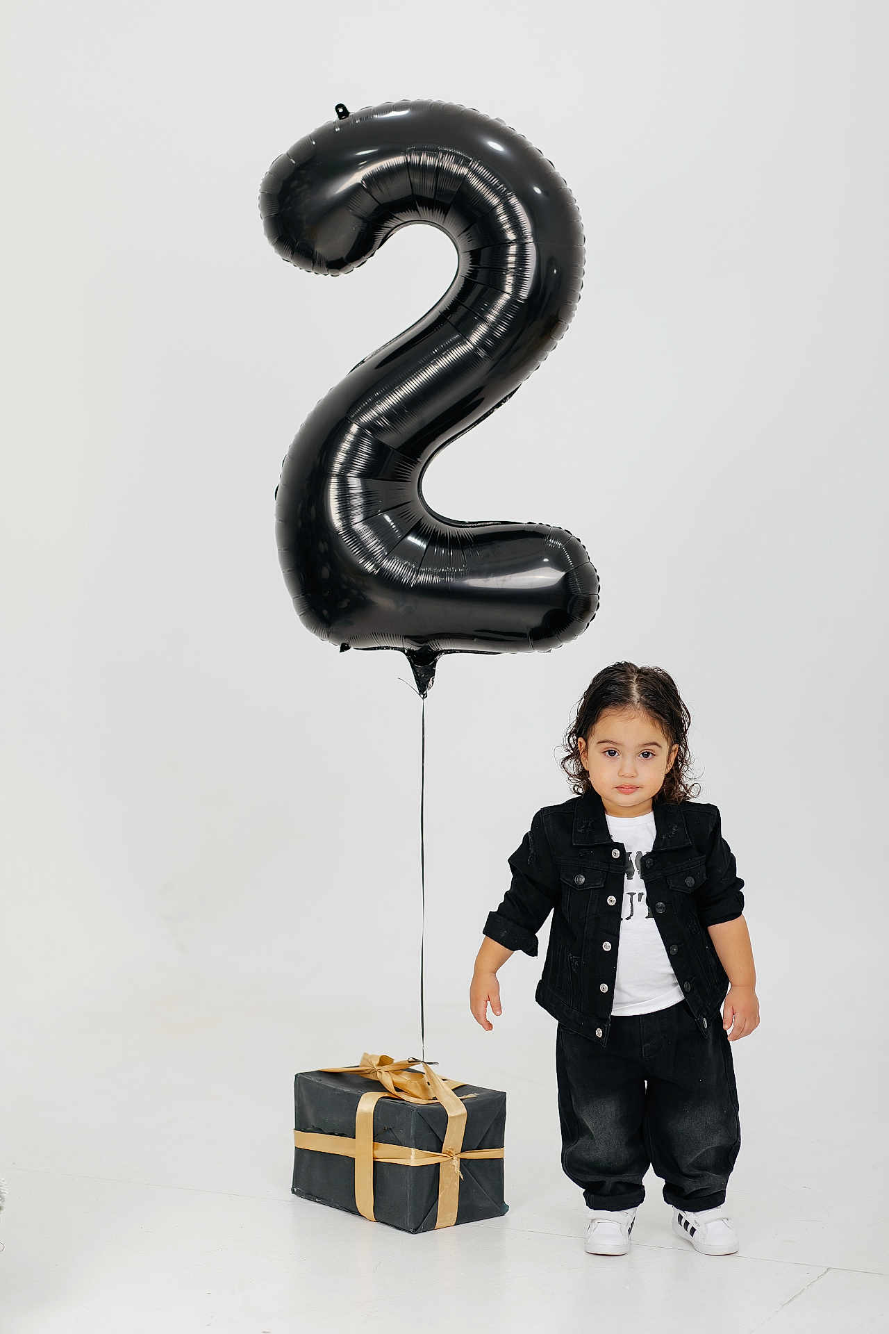 Ali is registered to the contest to win money with this photo: toddler, child, balloon, number_two, gift_box, black_clothing, white_shoes, studio, portrait, celebration, present, ribbon, standing, casual_wear, expressionless, indoor, minimalist, curly_hair, fashion, isolated