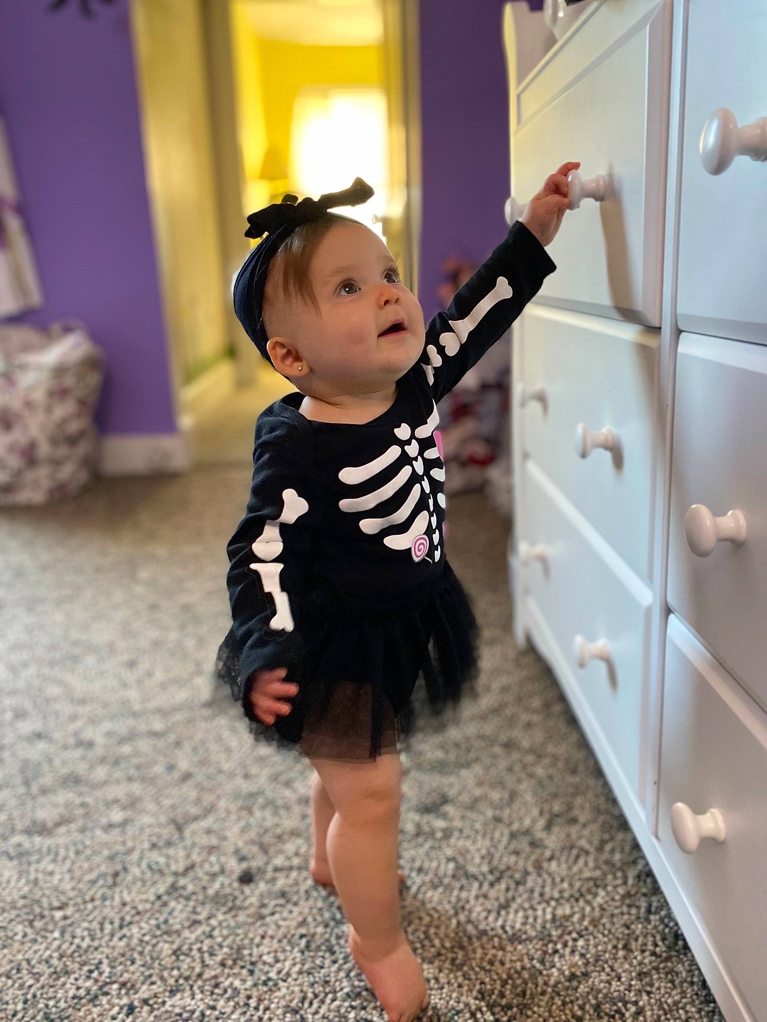 Veronica is registered to the contest to win money with this photo: baby, baby_toddler_clothing, cabinetry, day_dress, drawer, dresser, flooring, formal_wear, fun, happy, human_leg, one_piece_garment, pattern, person, purple, shoulder, sleeve, standing, toddler, waist