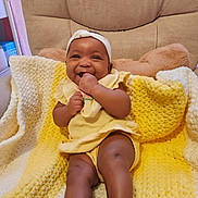 Naomi is registered to the contest to win money with this photo: baby, smiling, yellow_clothing, blanket, knit_blanket, infant, cute, happy, sitting, headband, hand, furniture, cushion, indoors, portrait, child, cozy, warm_colors, playful, young_child