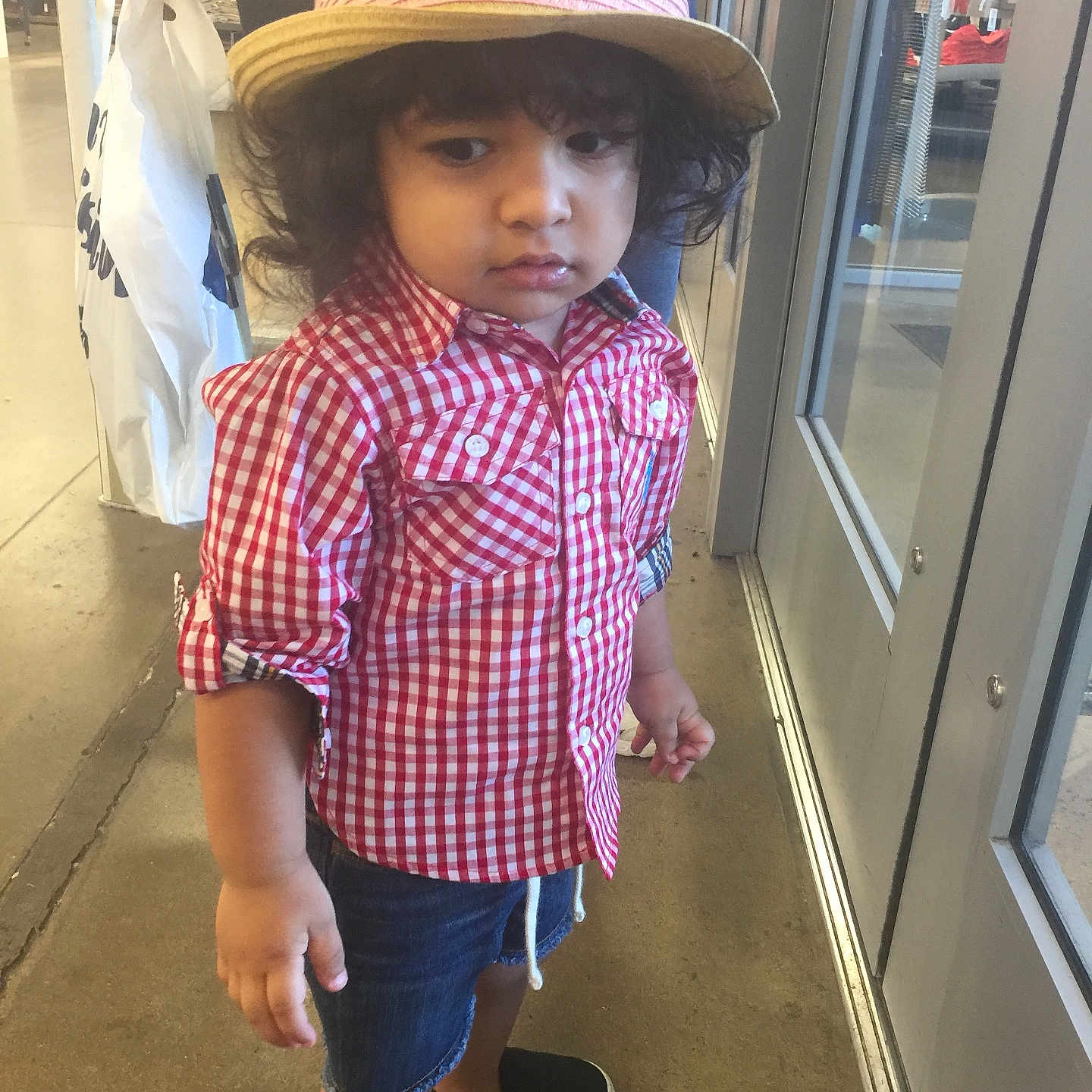Phelopateer is registered to the contest to win money with this photo: black_shoes, casual_clothing, child, curious_expression, curly_hair, denim_shorts, floor, glass_door, hand, hat, indoor, natural_light, person, red_checkered_shirt, retail_store, shopping_bag, standing, toddler, window, young_child