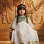 child, ancient_egypt, costume, dress, hieroglyphs, stool, portrait, indoor, historical_background, decorative, young_girl, headband, braided_hair, floor, wall_art, traditional, seated, cultural, photography, costume_party