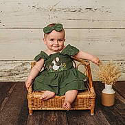Blair is registered to the contest to win money with this photo: baby, child, green_dress, bow, wicker_chair, smile, vase, dried_flowers, wooden_floor, wooden_wall, cute, sitting, portrait, indoor, infant, happy, person, small_furniture, decor, playful