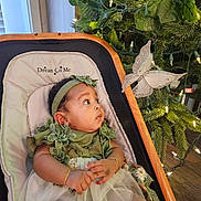 Angel joined the competition — help win amazing prizes! baby, infant, child, bassinet, green_clothing, headband, bracelet, butterfly_ornament, christmas_tree, lights, decorations, holiday, wooden_floor, indoor, curtains, window, face, expression, looking, cute