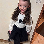 Joyce a rejoint le concours — aidez-le/la à gagner de superbes lots ! child, girl, smiling, curly_hair, bow, white_top, black_skirt, tights, standing, indoor, wall, light_switch, tile_floor, patterned_floor, person, happy, cute, fashion, portrait, flooring