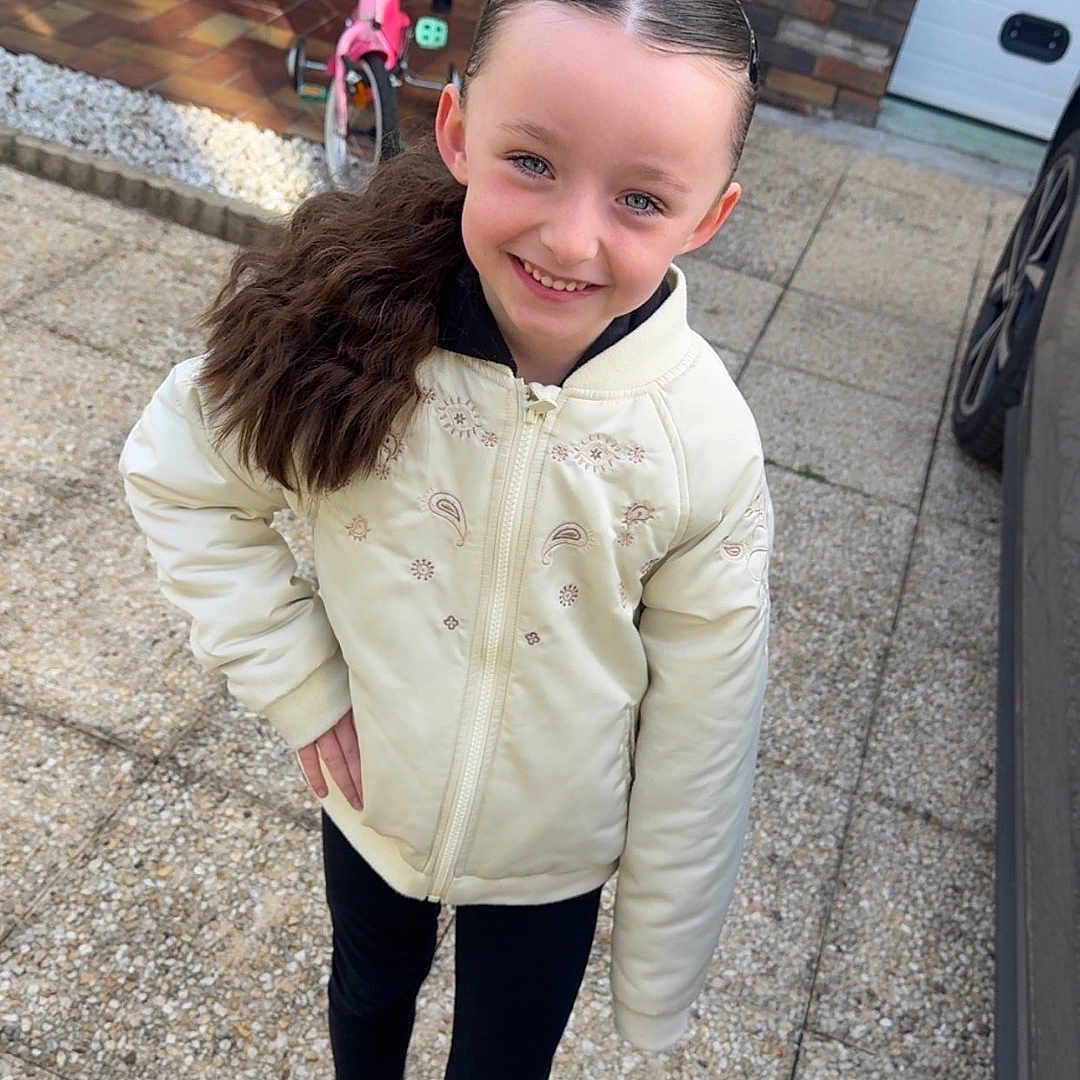 Joyce a rejoint le concours — aidez-le/la à gagner de superbes lots ! child, girl, smiling, outdoor, pavement, jacket, black_pants, sneakers, tricycle, toy, table, wooden_table, plant, hair_ponytail, happy, daylight, face, person, casual_clothing, backyard