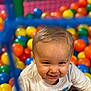child, baby, toddler, ball_pit, plastic_balls, smiling, portrait, indoor, play, colorful, short_hair, cheeky_grin, closeup, background_balls, kids_play_area, eye_contact, soft_lighting, fun, casual_clothing, netting