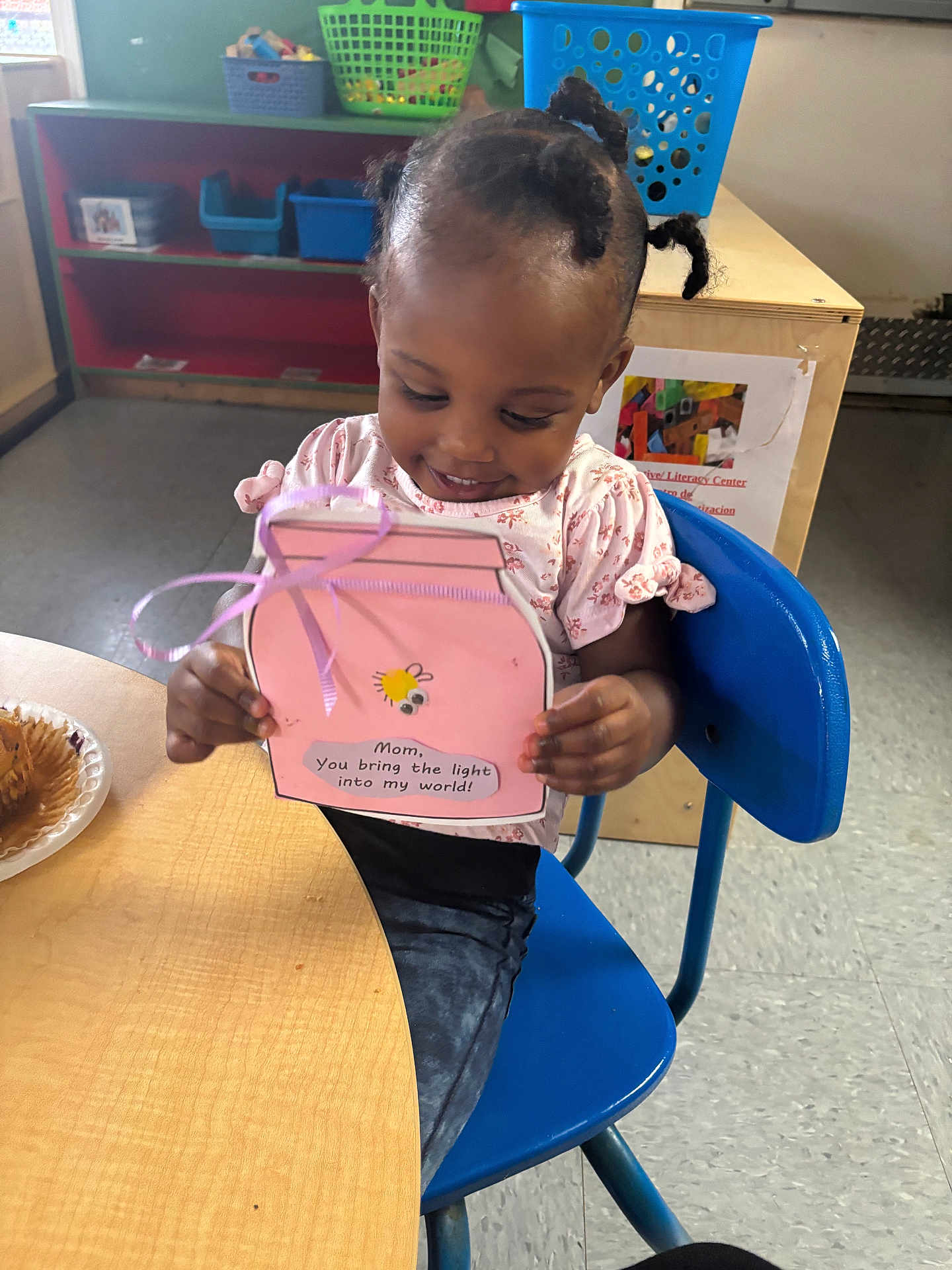 Jhazire joined the competition — help win amazing prizes! toddler, child, smiling, indoor, blue_chair, table, handmade_card, craft, bee_drawing, pink, clothing, hair, classroom, shelves, baskets, floor, plate, muffin, person, happy