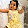 baby, child, smiling, yellow_outfit, butterfly_patch, hair_clip, cushion, face, cute, infant, happy, indoors, lying_down, arm_up, bracelet, black_hair, portrait, soft_lighting, baby_clothes, white_background