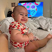Aleia is registered to the contest to win money with this photo: baby, smiling, diaper, strawberry_pattern, plush_toy, bed, indoor, television, child, cute, happy, person, clothing, furniture, toy, head, skin, face, room, home