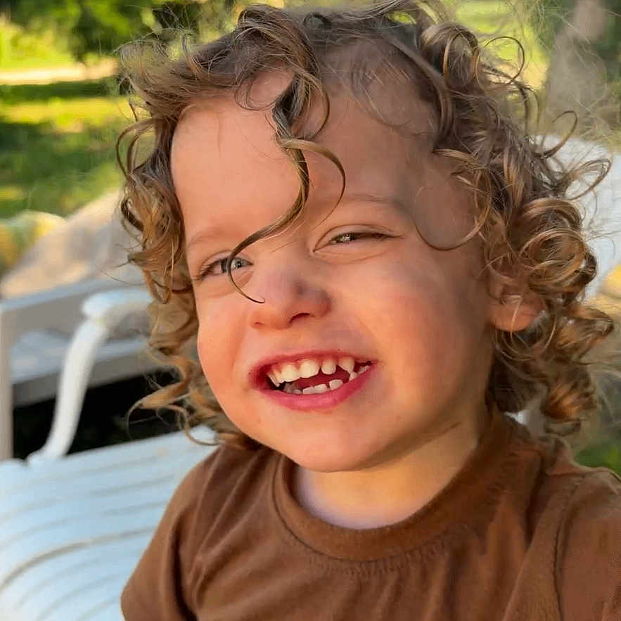 Kaïden participe au concours pour gagner de l'argent avec cette photo : child, toddler, curly_hair, smiling, outdoor, greenery, sunlight, brown_shirt, giraffe_print, white_chair, happy, portrait, nature, daylight, cute, person, grass, casual_clothing, young, playful