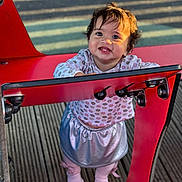 Zohra is registered to the contest to win money with this photo: toddler, child, smile, happy, playground, red_structure, outdoor, wooden_floor, clothing, skirt, long_sleeve, flower_pattern, shoes, legs, face, hair, sunlight, shadow, person, cute