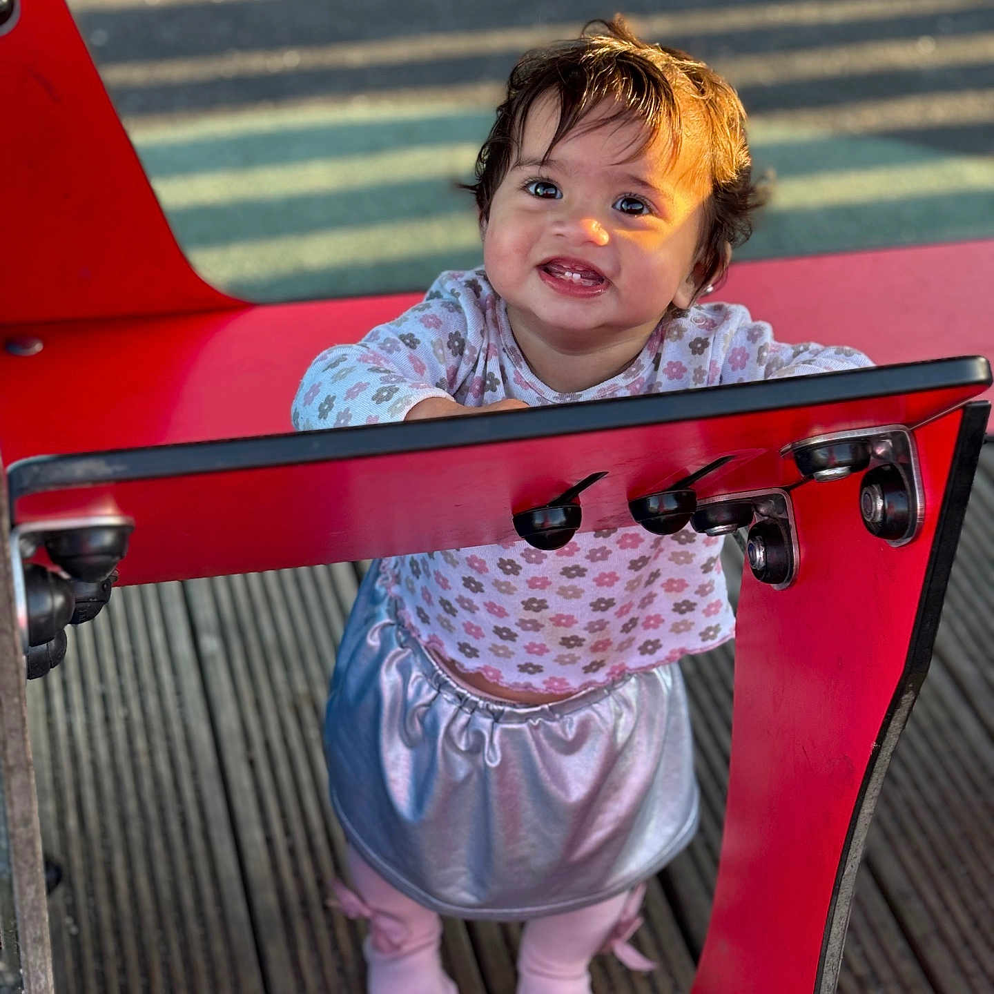 Zohra is registered to the contest to win money with this photo: child, clothing, cute, face, flower_pattern, hair, happy, legs, long_sleeve, outdoor, person, playground, red_structure, shadow, shoes, skirt, smile, sunlight, toddler, wooden_floor