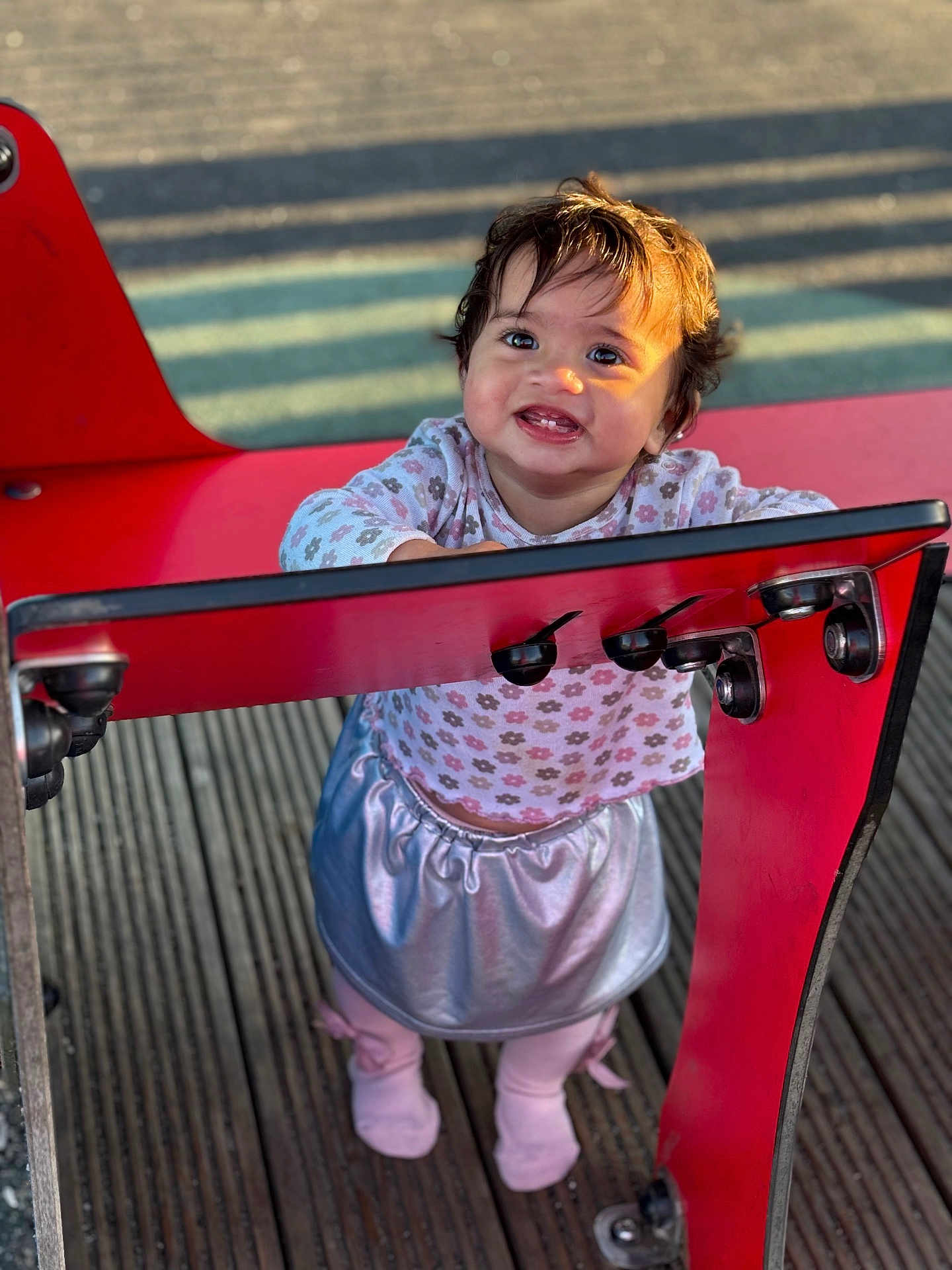 Zohra is registered to the contest to win money with this photo: toddler, child, smile, happy, playground, red_structure, outdoor, wooden_floor, clothing, skirt, long_sleeve, flower_pattern, shoes, legs, face, hair, sunlight, shadow, person, cute